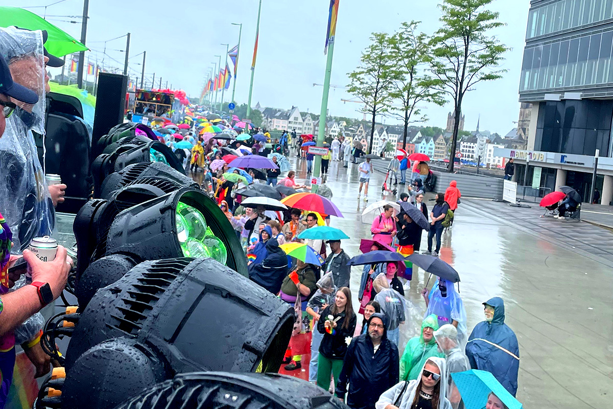 Zu sehen ist der Blick vom Festwagen der Firma müllermusic während der CSD-Parade in Köln. Trotz Regens stehen viele Menschen auf der Straße. Auf der linken Seite sind mehrere Scheinwerfer (Moving Lights) zu erkennen, die zur Veranstaltungstechnik auf dem Truck gehören.